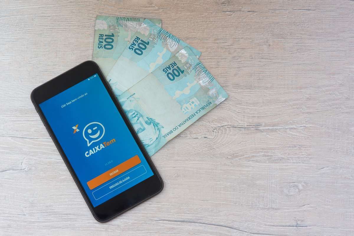 Um aplicativo Caixa Tem aberto no celular em cima de algumas notas de R$100,00. Veja aqui como conseguir empréstimo pelo Caixa Tem