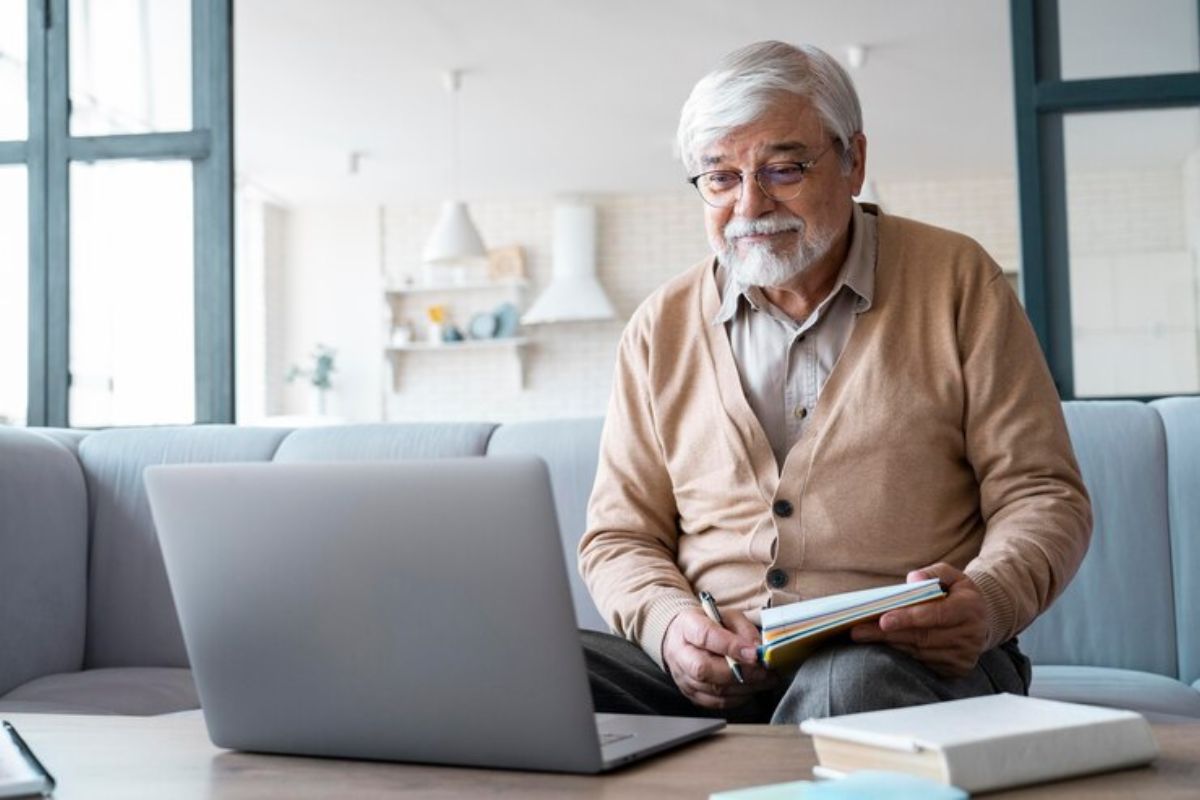 Um senhor na sala de sua casa com um caderno no colo e um notebook onde procura vagas de emprego para idosos